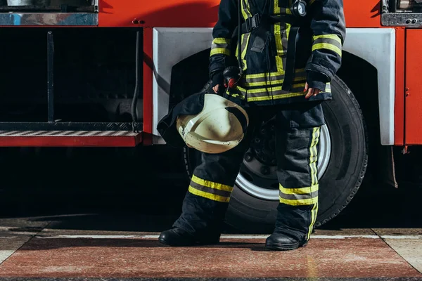 Partial view of female firefighter in protective uniform with helmet in hand standing near truck at fire station — Stock Photo