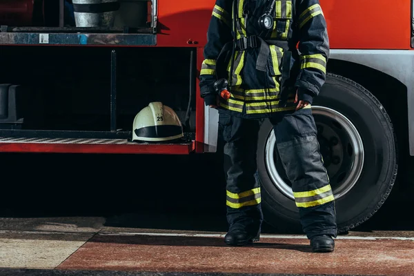 Vista parziale del pompiere donna in uniforme protettiva in piedi vicino camion con casco alla stazione dei pompieri — Foto stock