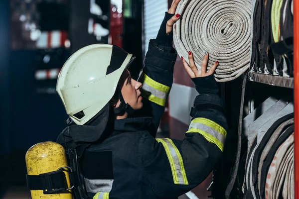 Vista laterale del vigile del fuoco femminile mettendo tubo dell'acqua nel camion alla stazione dei pompieri — Foto stock