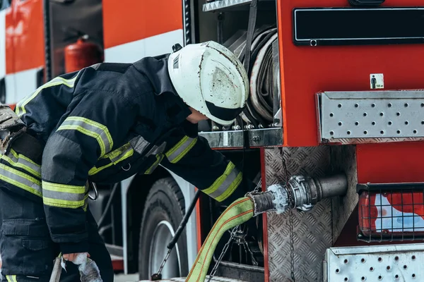 Firefighter in protective uniform checking water hose in truck on street — Stock Photo