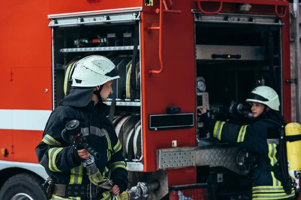 Pompier masculin en uniforme tenant tuyau d'eau tandis que collègue vérifier l'équipement dans le camion sur la rue — Photo de stock