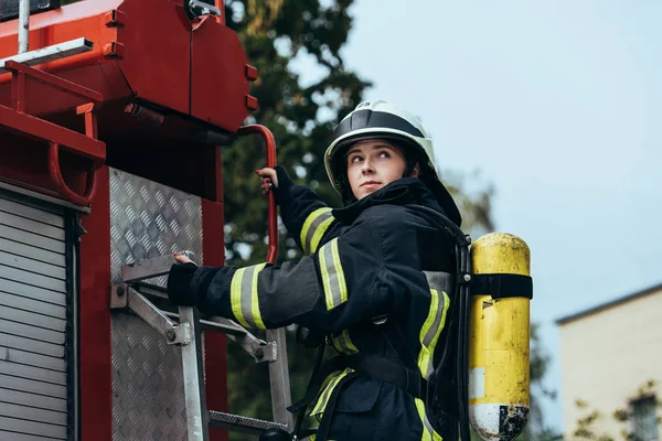 Female firefighter with fire extinguisher on back standing on fire truck on street — Stock Photo