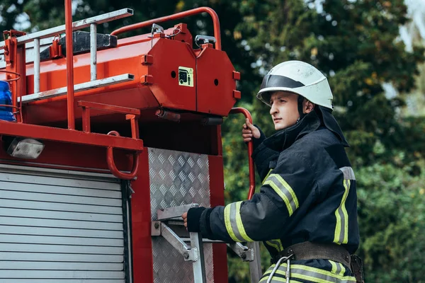 Male firefighter in protective helmet standing on truck on street — Stock Photo