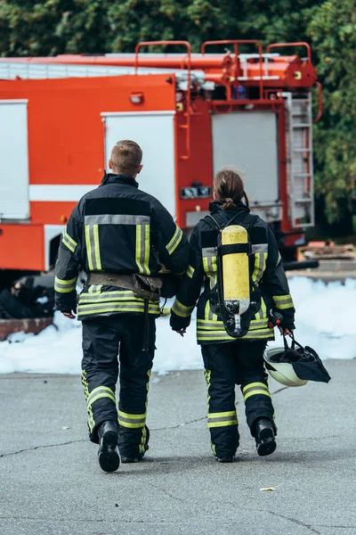 Vista trasera de los bomberos en uniforme ignífugo caminando por la calle con camión de bomberos detrás - foto de stock