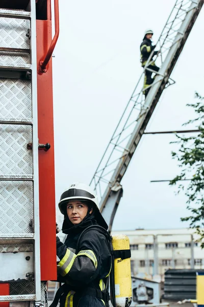 Bombeiro feminino em uniforme e capacete olhando para longe, enquanto colega de pé na escada na rua — Fotografia de Stock