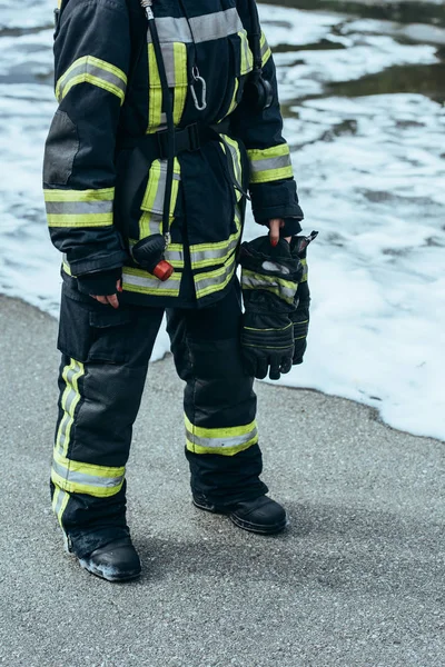Vue partielle de la femme pompier en uniforme ignifuge avec des gants à la main debout sur le steet avec de la mousse — Photo de stock