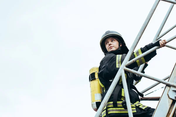 Femme pompier en uniforme de protection et casque avec extincteur sur le dos debout sur l'échelle avec ciel bleu sur fond — Photo de stock