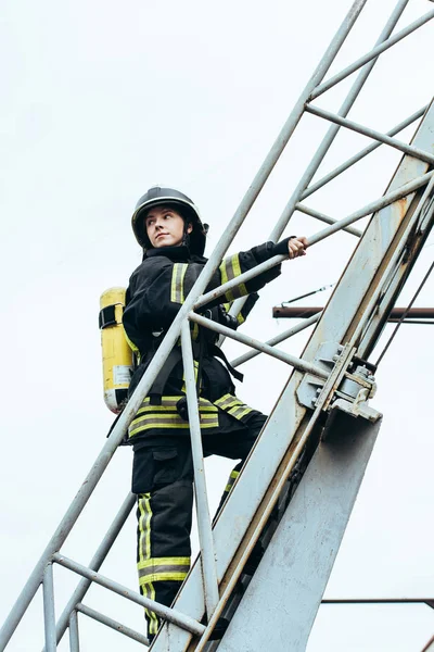 Bombero femenino en uniforme protector y casco con extintor de incendios en la espalda de pie en escalera con cielo azul en el fondo - foto de stock