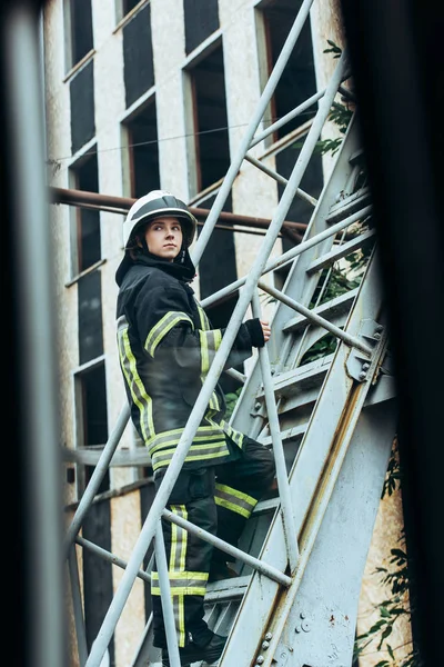 Пожарная женщина в защитной форме и шлеме стоит на лестнице на улице — стоковое фото