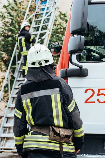 Rückansicht eines Feuerwehrmannes in Schutzuniform und Helm, der seinen Kollegen auf der Leiter auf der Straße ansieht — Stockfoto