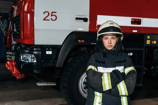 Портрет жінки пожежника в шоломі з обіймами, схрещеними стоячи на пожежній станції з вантажівкою позаду — стокове фото