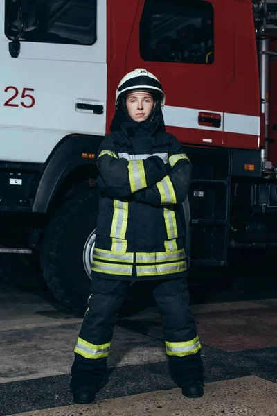 Feuerwehrfrau im Helm mit gekreuzten Armen steht am Feuerwehrhaus mit LKW dahinter — Stockfoto