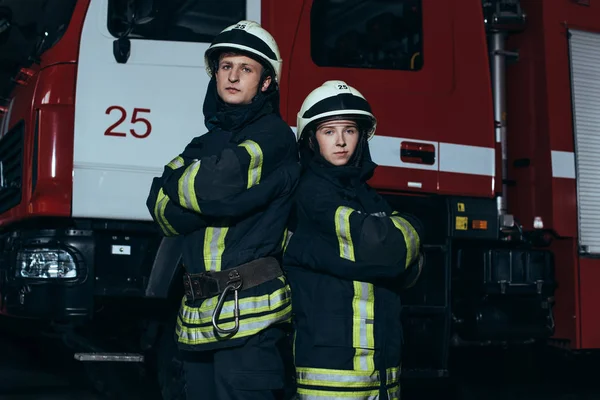 Retrato de bomberos en uniforme ignífugo y cascos con los brazos cruzados de pie cerca del camión en la estación de bomberos - foto de stock