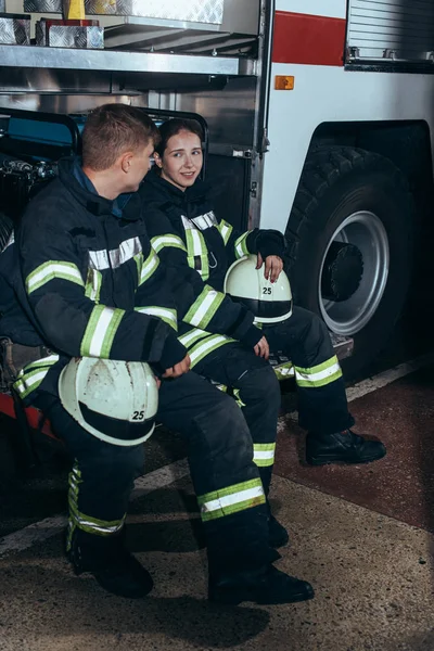 Bomberos en uniforme de protección teniendo conversación cerca de camión en estación de bomberos - foto de stock