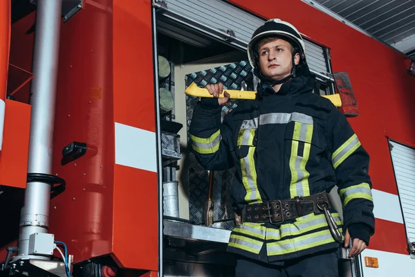 Vista a basso angolo del vigile del fuoco in casco con ascia guardando lontano alla stazione dei pompieri — Foto stock
