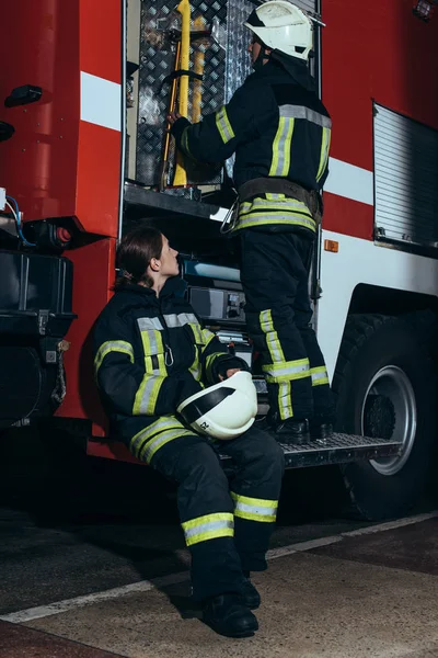 Femme pompier en uniforme de protection regardant collègue vérifier l'équipement dans le camion au service d'incendie — Photo de stock