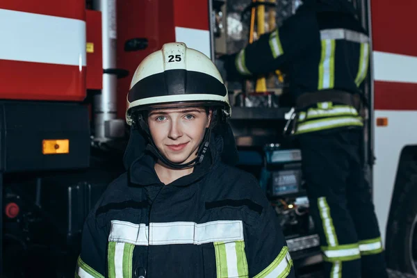 Femme pompier souriante dans le casque regardant la caméra avec un collègue vérifier l'équipement derrière à la caserne de pompiers — Photo de stock