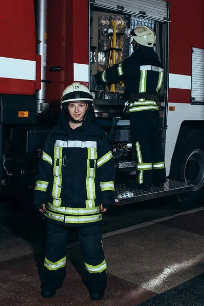 Lächelnde Feuerwehrfrau blickt in Kamera, während Kollegin Ausrüstung im LKW bei der Feuerwehr überprüft — Stockfoto