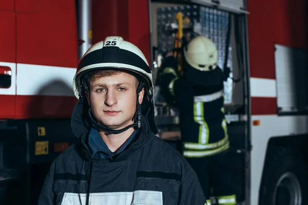 Male firefighter in helmet looking at camera with colleague checking equipment behind at fire station — Stock Photo