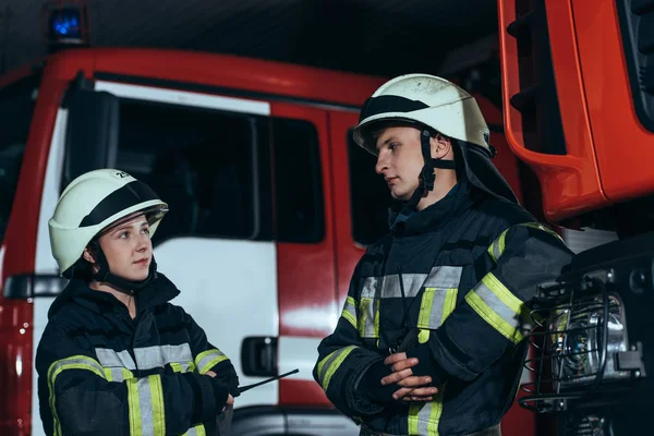 Пожарные в огнеупорной форме и шлемах, смотрящие друг на друга на пожарной станции — стоковое фото