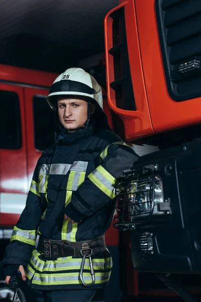 Porträt eines Feuerwehrmannes in Uniform und Helm, der sich am Feuerwehrhaus auf einen Lastwagen stützt — Stockfoto