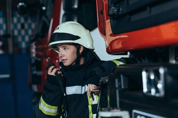 Retrato de uma mulher bombeira de uniforme protector a falar em rádio portátil no quartel dos bombeiros — Stock Photo