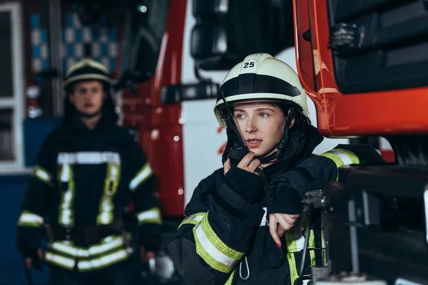 Fuoco selettivo di pompiere donna che parla su apparecchio radio portatile con collega dietro a pompieri — Foto stock