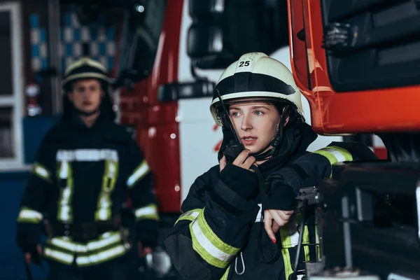 Foyer sélectif des femmes pompiers parlant sur un poste de radio portable avec un collègue derrière au service d'incendie — Photo de stock