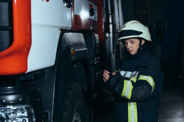 Bombero femenino con casco comprobando el uniforme protector en la estación de bomberos - foto de stock
