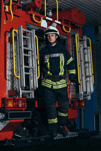 Fireman in protective uniform and helmet standing on truck at fire station — Stock Photo