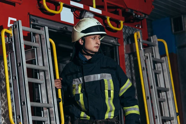 Bombero con uniforme protector y casco mirando a la estación de bomberos - foto de stock