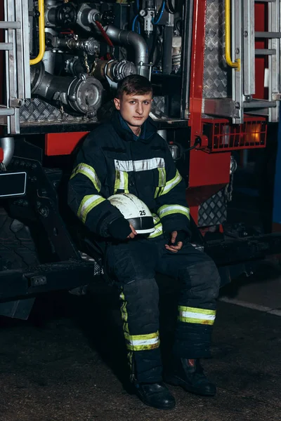 Maschio vigile del fuoco in uniforme con casco guardando la fotocamera al camion alla stazione dei pompieri — Foto stock