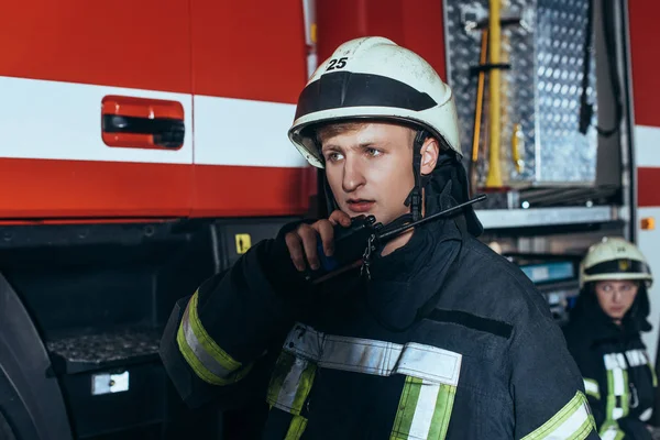 Foyer sélectif des pompiers en uniforme parlant sur un poste radio portable avec un collègue derrière à la caserne de pompiers — Photo de stock