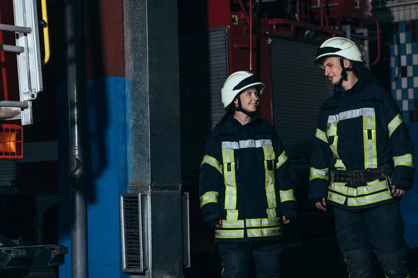 Bomberos sonrientes con uniforme protector y cascos mirándose en la estación de bomberos - foto de stock