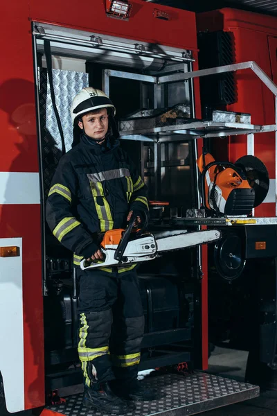 Bombero en casco sosteniendo la sierra eléctrica mientras está parado en el camión en la estación de bomberos — Stock Photo