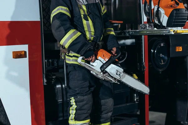 Vista parcial del bombero en uniforme protector sosteniendo la sierra eléctrica en las manos en la estación de bomberos - foto de stock