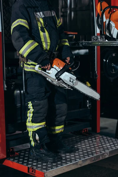 Visão parcial de bombeiro em uniforme de proteção segurando serra elétrica em mãos no quartel de bombeiros — Fotografia de Stock