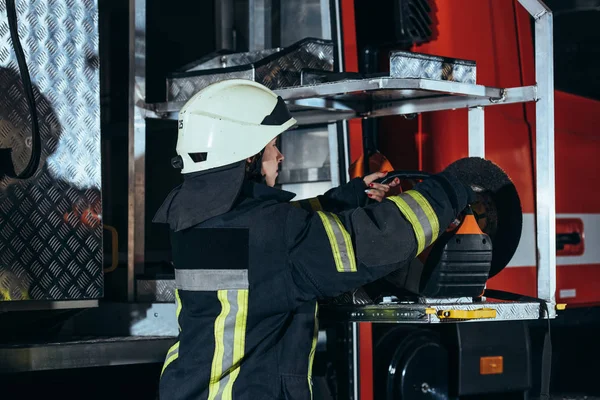 Vista laterale del vigile del fuoco femminile in piedi su attrezzature in camion alla stazione dei pompieri — Foto stock