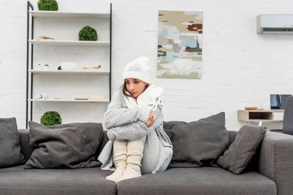Erfrorene junge Frau in warmer Kleidung, die zu Hause auf der Couch sitzt und wegschaut — Stockfoto