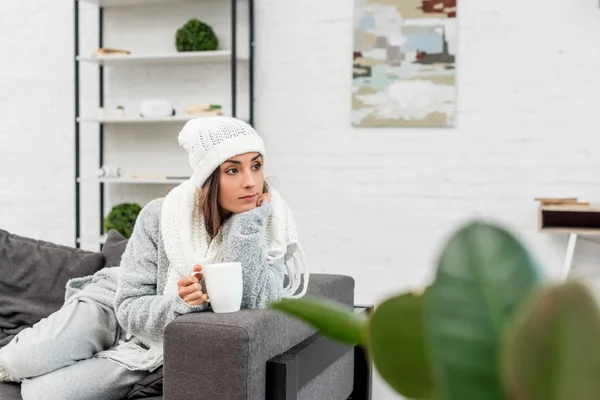 Вдумлива молода жінка в теплому одязі тримає чашку гарячого чаю і дивиться геть, сидячи на дивані — стокове фото
