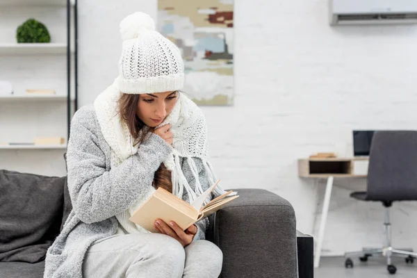 Сфокусированная молодая женщина в теплой одежде сидит дома на диване и читает книгу — стоковое фото