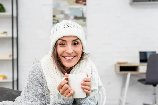 Sorrindo jovem mulher em roupas quentes segurando xícara de chá quente em casa — Fotografia de Stock