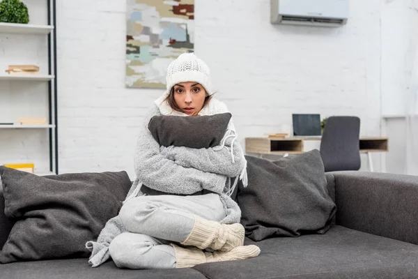 Заморожена молода жінка в теплому одязі сидить на дивані і обіймає подушку вдома — стокове фото
