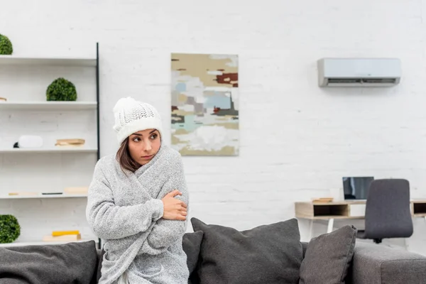 Mujer joven congelada en ropa de abrigo sentada en el sofá en casa con aire acondicionado colgando en el fondo - foto de stock