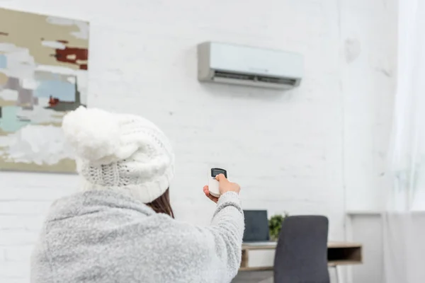 Rear view of freezed young woman in sweater pointing at air conditioner with remote control — Stock Photo