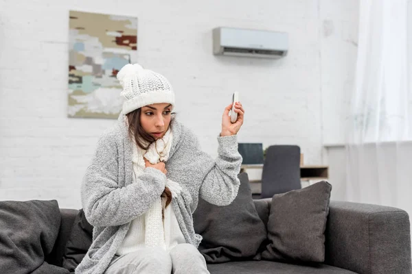 Freezed young woman in sweater pointing at air conditioner with remote control — Stock Photo