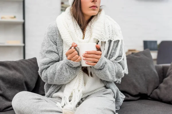 Abgeschnittene Aufnahme einer kranken jungen Frau in warmer Kleidung, die eine Tasse Heißgetränk hält, während sie zu Hause auf der Couch sitzt — Stockfoto