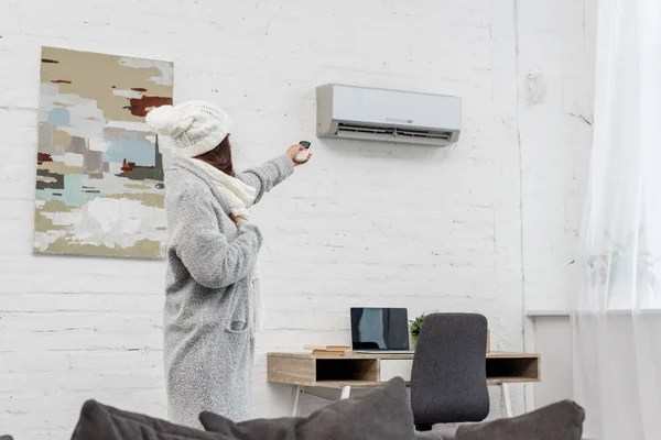 Schöne junge Frau in warmer Kleidung zeigt auf Klimaanlage mit Fernbedienung zu Hause — Stockfoto