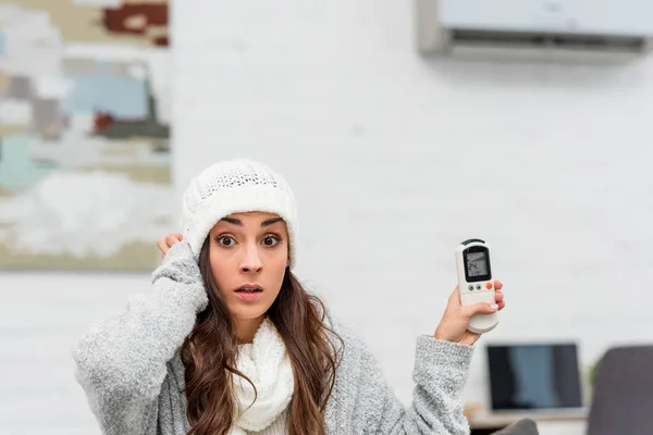 Shocked young woman in warm clothes holding air conditioner remote control at home — Stock Photo