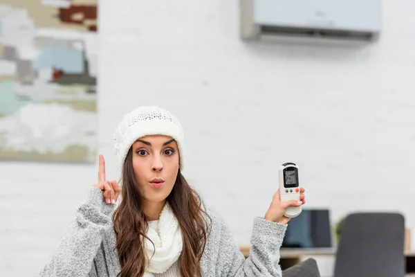 Congelé jeune femme en vêtements chauds ayant idée tout en tenant climatiseur télécommande à la maison — Photo de stock